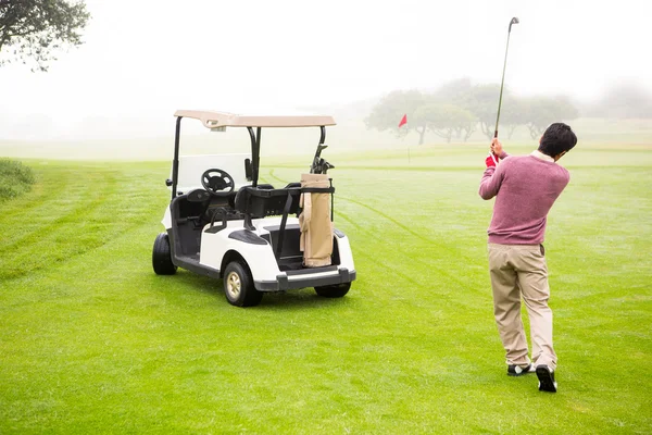 Golfer teeing off next to his golf buggy