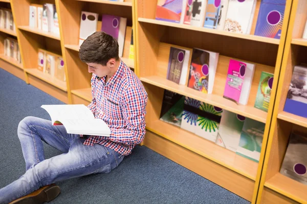 Young man reading book while sitting by bookshelf