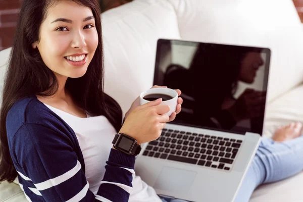 Asian woman relaxing on couch with coffee using laptop