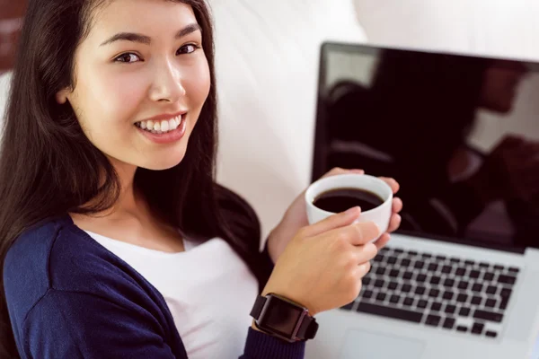 Asian woman relaxing on couch with coffee using laptop