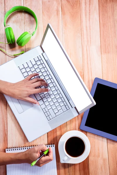 Overhead of feminine hand typing on laptop and taking notes