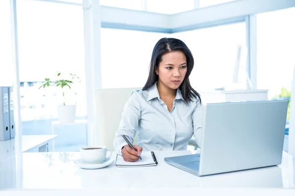 Serious businesswoman writing on notepad and using laptop