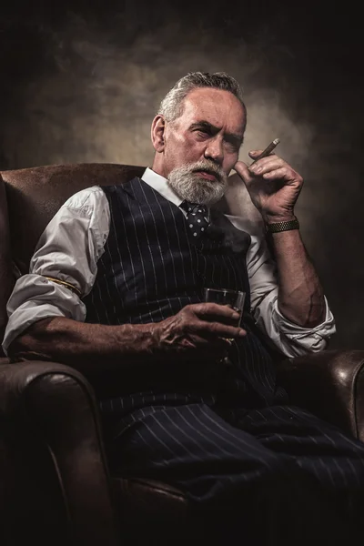 In chair sitting senior business man with cigar and whisky. Gray