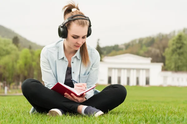 Young beautiful female sitting and writing outside with earphone