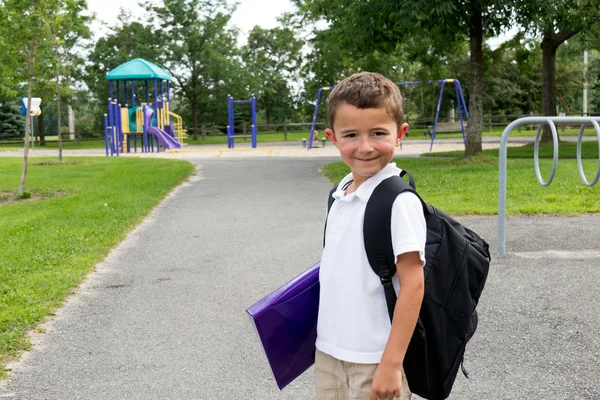 Happy little boy with school backpack and book in the play yard