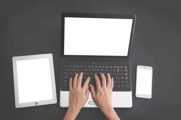 Hands typing on laptop with tablet and mobile phone on the side.