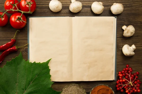 Open old vintage book with berries, tomatoes, Chile peppers, spices and grape leaf on wooden background. Healthy vegetarian food. Recipe, menu, mock up, cooking.