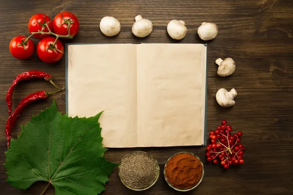 Open old vintage book with berries, tomatoes, Chile peppers, spices and grape leaf on wooden background. Healthy vegetarian food. Recipe, menu, mock up, cooking.