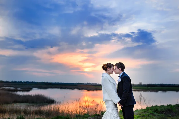 Portrait of beautiful bride and groom hugging and looking at lake at sunset
