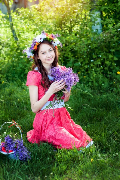 Beautiful woman in red dress sitting on the grass with a branch of lilac