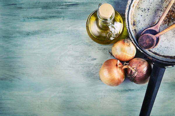 Old kitchen pan wooden spoon three onions carafe with olive oil on wooden table.