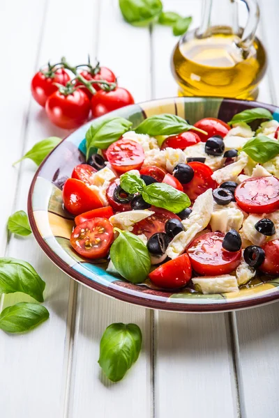 Caprese. Caprese salad. Italian salad. Mediterranean salad. Italian cuisine. Mediterranean cuisine. Tomato mozzarella basil leaves black olives and olive oil on wooden table.