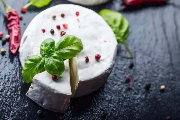 Fresh Brie cheese and a slice on a granite board with basil leaves four colors peper and chili pepers. Italian and Mediterranean ingredients.