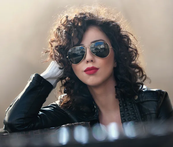 Charming young curly brunette woman with sunglasses and black leather jacket against wall. Sexy gorgeous young woman with modern look. Portrait of sensual girl with voluptuous mouth