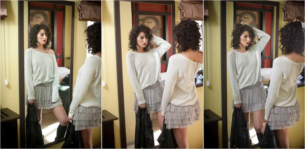 Young woman in white blouse and gray short tutu skirt looking into large mirror holding black leather jacket. Beautiful curly dark hair girl posing in front of wall mirror. Fashionable brunette model.