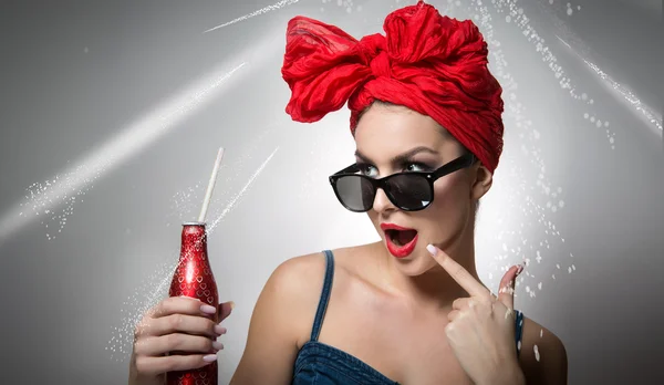 Woman with red turban and sunglasses holding a drink bottle with a straw inside. Attractive girl portrait with bottle in hand, studio shot on gray background. Happy young female, advertisement concept