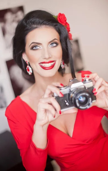 Beautiful young woman with creative make-up and hair style taking photos with a camera. Fashionable attractive brunette with Spanish look holding a camera. Lady in red with flower in hair, smiling