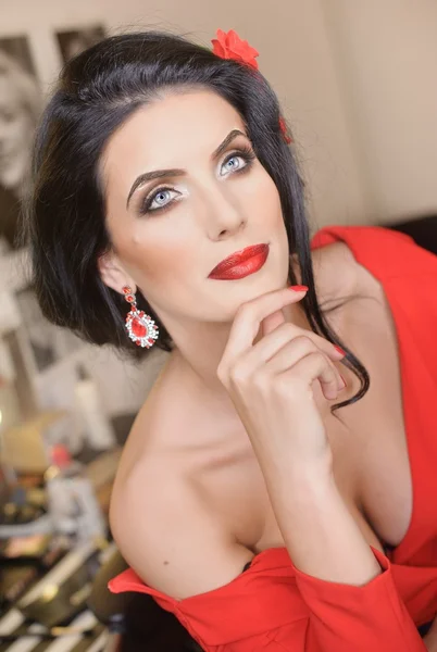 Beautiful young woman with creative make-up and hair style posing. Fashionable attractive brunette with Spanish look, indoors shot. Portrait of lady in red with flower in hair and gorgeous eyes