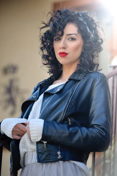 Charming young curly brunette woman with black leather jacket. Sexy gorgeous young woman with modern look. Portrait of sensual girl with voluptuous mouth wearing bright red lipstick, outdoor shot