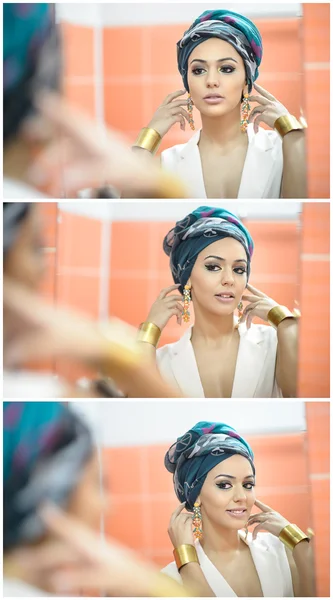 Young beautiful woman with turban and golden accessories looking into large mirror. Seductive lady with earrings posing, mirror reflection photo. Elegant girl in white smiling posing into mirror