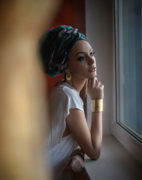 Attractive sexy lady in white blouse posing in window frame looking outside. Portrait of sensual young woman with turban. Gorgeous female with beautiful eyes daydreaming enjoying the bright day light