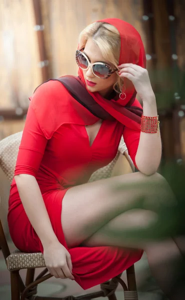 Fashionable attractive lady with red dress and headscarf sitting on chair in restaurant, outdoor shot in sunny day. Young short hair blonde woman with sunglasses posing outside exposing legs