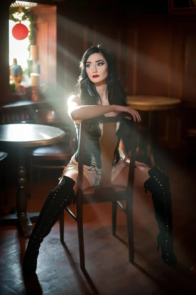 Beautiful sexy girl with long leather boots sitting on chair in comfortable position. Brunette woman posing challenging. Sensual female with black corset and high heels in vintage scenery