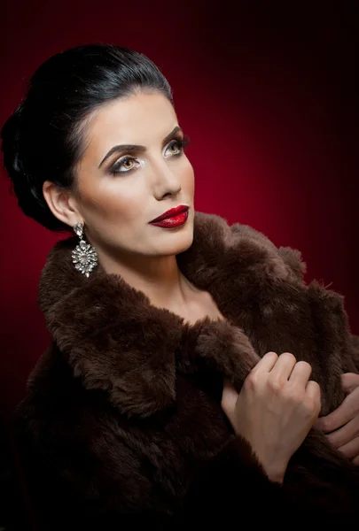 Attractive sexy young woman wearing a fur coat posing in studio on dark purple background. Portrait of sensual female with creative makeup, studio. Gorgeous eyes brunette lady with black fur collar
