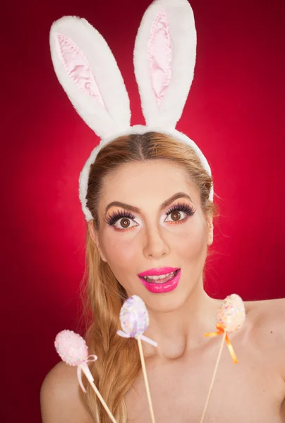 Beautiful blond woman as Easter bunny with rabbit ears on red background, studio shot. Young lady holding three pastel colored eggs lollipops, Easter concept. Beautiful eyes bunny girl portrait