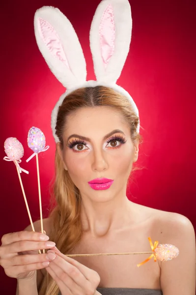 Beautiful blond woman as Easter bunny with rabbit ears on red background, studio shot. Young lady holding three pastel colored eggs lollipops, Easter concept. Beautiful eyes bunny girl portrait