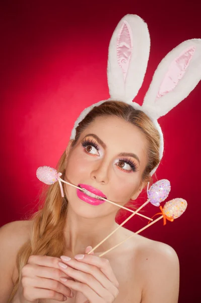 Beautiful blond woman as Easter bunny with rabbit ears on red background, studio shot. Young lady holding three pastel colored eggs lollipops, Easter concept. Beautiful eyes bunny girl portrait