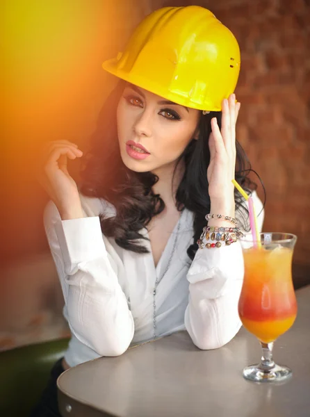 Beautiful woman civil engineer with yellow helmet taking a break in front of orange juice. Young female architect with white shirt drinking a juice in restaurant. Young female construction specialist