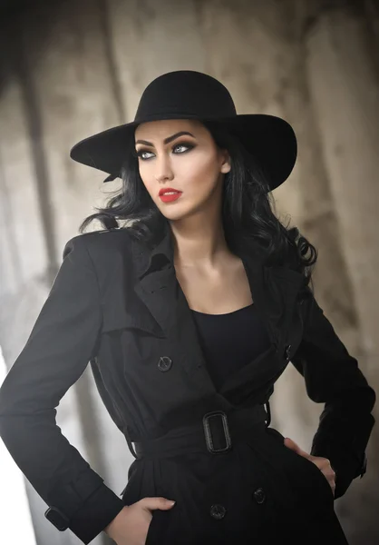 Beautiful young woman in black outfit, urban concept. Elegant brunette with hat posing with hands in her pocket, outdoor. Romantic girl with gorgeous blue eyes wearing black coat with buckle.