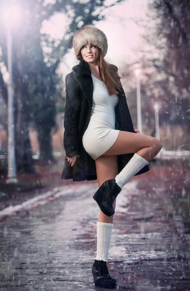 Portrait of young beautiful woman, outdoor shot in winter scenery. Sensual brunette girl with coat and fur cap posing in a park covered with snow. Fashionable female in a cold day.