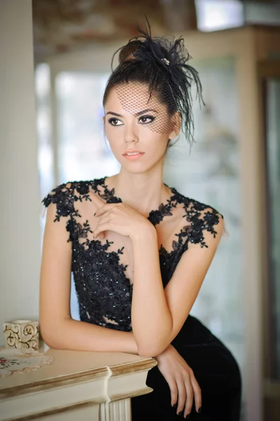 Beautiful brunette lady in elegant black lace dress posing in a vintage scene. Young sensual fashionable woman with creative hairstyle indoor. Attractive slim girl indoor shot in luxurious scenery