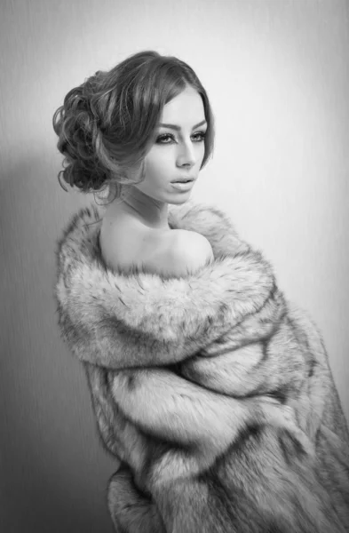 Attractive sexy young woman wearing a fur coat posing provocatively indoor. Portrait of sensual female with creative haircut, studio shot. Beautiful girl covered only with a fur exposing her shoulders