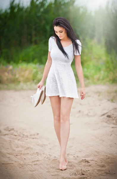 Attractive brunette girl with short white dress strolling barefoot on the countryside road. Young beautiful woman walking with shoes in hand with forest in background. Female with long legs outdoor