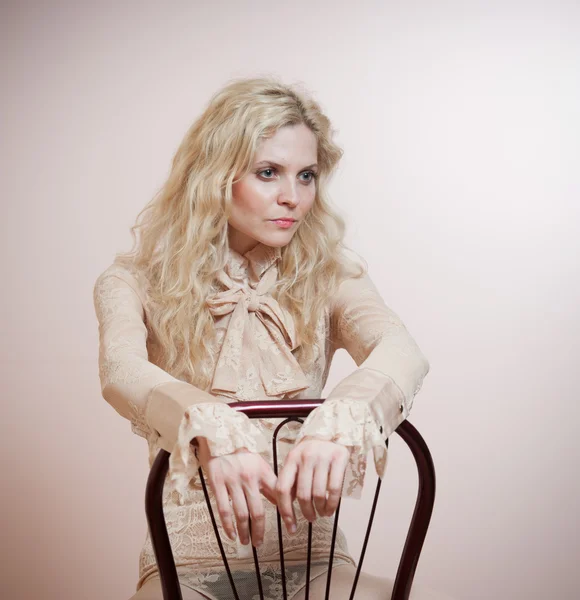 Attractive fair hair model with in elegant nude blouse sitting provocatively on chair, studio shot. Fashion portrait of a sensual blonde woman in classic blouse with long sleeves and ribbon on chair