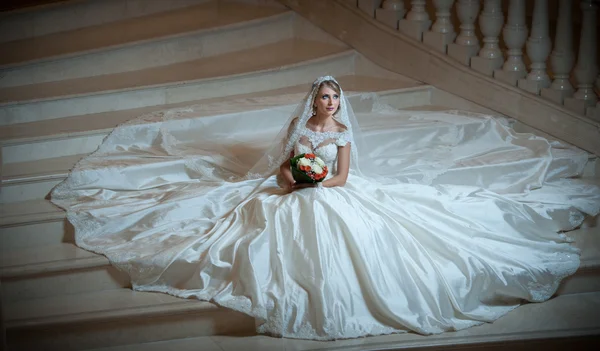 Young beautiful luxurious woman in wedding dress sitting on stair steps in semi-darkness. Bride with huge wedding dress holding her bouquet. Seductive blonde bride with gorgeous gown posing