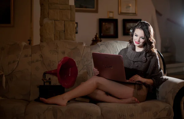 Beautiful young woman sitting on sofa working on laptop having a red gramophone near her, in boudoir scenery. Attractive brunette girl with long hair and long legs laying down on couch with a laptop