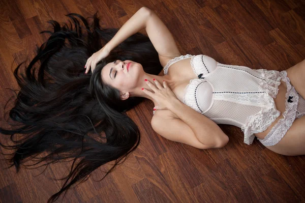 Beautiful sexy brunette young woman wearing white lingerie lying on wooden floor. Sensual gorgeous perfect body female posing provocatively, boudoir shot indoor. Attractive long hair girl in corset.
