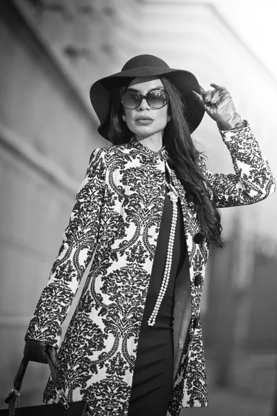 Attractive young woman in a winter fashion shot. Beautiful fashionable young girl in black posing on avenue. Elegant brunette with hat, sunglasses and handbag in urban scenery.