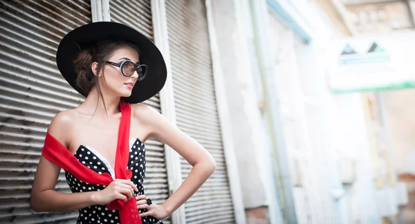 Attractive brunette girl with black hat, red scarf and sunglasses posing outdoor. Beautiful fashionable young woman with modern accessories, urban shot. Gorgeous brunette with large black hat smiling.