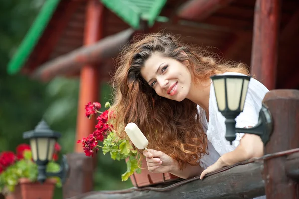 Beautiful female portrait with long brown hair eating ice cream near a pot with red flowers outdoor. Attractive woman with beautiful eyes smiling enjoying an ice cream in a summer day, outdoor shot