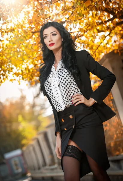 Attractive young woman in a autumnal fashion shot. Beautiful fashionable lady in black and white outfit posing provocatively in park. Elegant brunette in fall scenery on faded trees background .