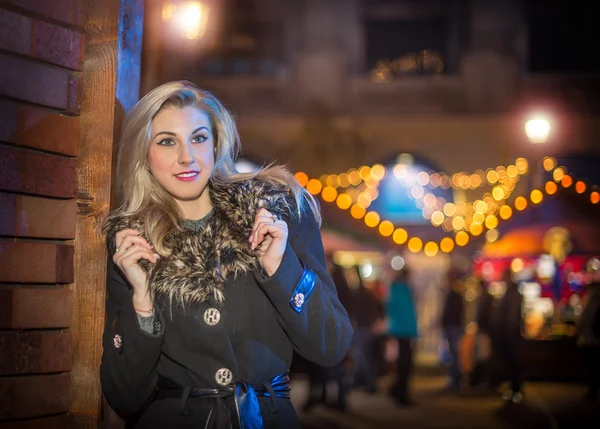Portrait of young beautiful woman with long fair hair outdoor in cold winter evening. Beautiful blonde girl in winter clothes with xmas lights in background. Beautiful woman smiling in winter scenery