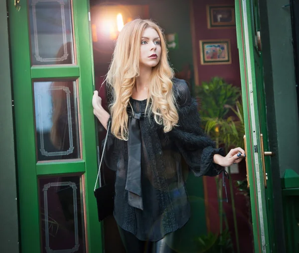 Charming young blonde woman in black outfit posing in a green painted door frame. Sexy gorgeous young woman with long curly hair opening the door for somebody. Portrait of sensual woman in black