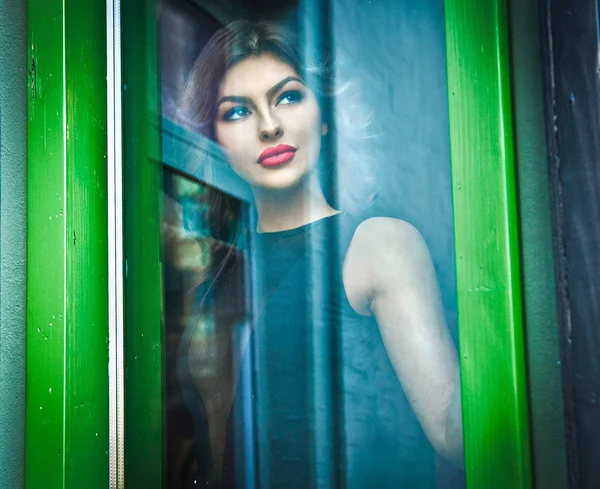 Beautiful sexy woman posing in a green painted window frame, shot through window. Sexy gorgeous young female with long curly hair looking outdoor for somebody. Portrait of sensual girl in black blouse