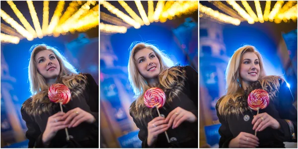 Portrait of young woman with long fair hair posing outdoor in cold winter evening. Beautiful blonde holding a big lollipop with Xmas lights on background. Attractive female smiling in Xmas market