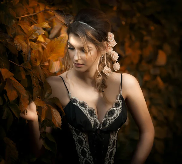Beautiful sensual woman with roses in hair posing near a wall of green leaves. Young female in black elegant dress daydreaming in nature. Attractive voluptuous lady with creative hair arrangement